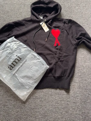 Svart hoodie med röd Ami-dekal - Säljer en svart hoodie från Ami med stor röd hjärt-A patch på bröstet. Hoodien har huva med dragsko och ribbade muddar vid ärmslut och nederkant. Materialet känns mjukt och skönt, perfekt för en avslappnad stil.