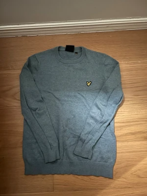 Blå tröja från Lyle & Scott - Snygg blå rundhalsad tröja från Lyle & Scott med den klassiska gula loggan broderad på bröstet. Tröjan har långa ärmar, ribbade muddar och är tillverkad i mjukt stickat material. Perfekt för en stilren och enkel look.
