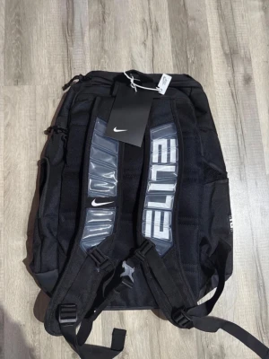 Svart Nike Elite ryggsäck - Snygg svart ryggsäck från Nike med ELITE-tryck på axelbanden och vita detaljer. Axelbanden har vadderade, genomskinliga partier och justerbara spännen. Ryggsäcken har flera fack och dragkedjor, perfekt för skola eller träning.