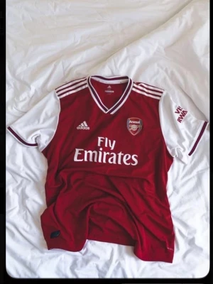 Arsenal matchtröja Adidas röd/vit - Säljer en Arsenal matchtröja från Adidas i rött och vitt med klubbmärke på bröstet och Fly Emirates-tryck. Tröjan har vita ärmar, röda detaljer och klassiska Adidas-ränder på axlarna. Perfekt för fotbollsfans!