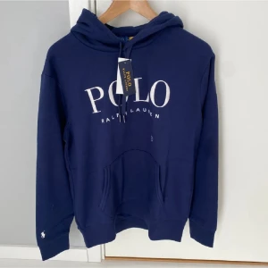Marinblå hoodie från Polo Ralph Lauren - Snygg marinblå hoodie från Polo Ralph Lauren med stor vit logga på bröstet och klassisk huva. Hoodien har känguruficka framtill och ribbade muddar vid ärmar och nederkant. Perfekt för en avslappnad och trendig look. Alla tags kvar!!! Helt oanvänd och är i bäst skick! pris går att pruta 
