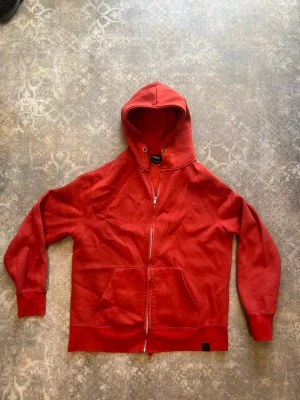 Röd hoodie med dragkedja Pull&Bear - Snygg röd hoodie från Pull&Bear med dragkedja framtill och två stora fickor.Sömmen har släppt lite i ena kanten på fickan. Hoodien har huva med metallöljetter och är gjord i mjukt bomullsmaterial med fleece på insidan. Perfekt för chill dagar eller när du vill ha en enkel streetstyle-look.
