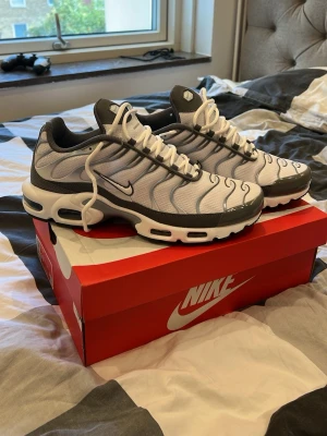 Nike Air Max Plus vita/grå sneakers - Nike Air Max Plus sneakers i vitt och grått med svarta detaljer. Skorna har en ikonisk vågig design, synlig Air-dämpning i sulan och mesh-överdel för bra andningsförmåga. Perfekta för dig som gillar streetwear och vill sticka ut med klassisk Nike-stil.