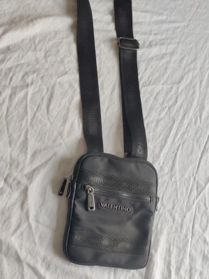 Svart axelväska från Valentino - Snygg svart axelväska från Valentino med justerbar axelrem och flera dragkedjefack. Väskan har en stilren, fyrkantig form och är tillverkad i ett slätt, tåligt material med diskret logga på både rem och framsida. Perfekt för att bära det viktigaste.