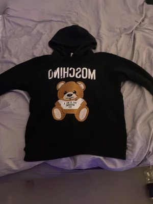 Svart hoodie med nalle från Moschino - Svart hoodie från Moschino med stor broderad nalle på bröstet och texten 'THIS IS NOT A MOSCHINO TOY'. Hoodien har långa ärmar och huva, samt en oversized passform. Perfekt för dig som gillar statement-plagg och lekfull design.