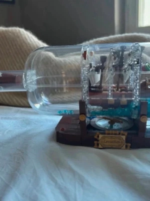 Lego skepp - Byggsats från Lego som föreställer ett skepp inuti en genomskinlig flaska med brun och guld dekor. Flaskan står på ett detaljerat ställ med kompass och skylt. Perfekt för dig som gillar unika samlarprylar och pyssel. och du kan få den ihop plockad om du vill bygga den själv!! Fattas en bit!!