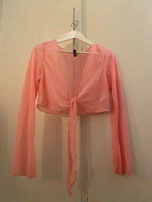 Rosa omlottopp från H&M Divided - Säljer en ljusrosa, transparent omlottopp från H&M Divided. Toppen har långa ärmar, croppad passform och knyt framtill. Perfekt för att styla lager på lager eller till ett par höga jeans.