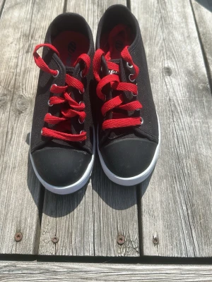 Svarta sneakers med röda skosnören - Snygga svarta sneakers med röda skosnören och vit sula. Skorna har en låg profil och är tillverkade i canvasmaterial med gummitå. Perfekta för en avslappnad och cool stil. Röd detalj på sidan ger extra edge.