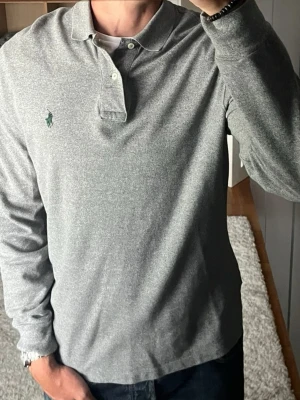 Polo Ralph Lauren långärmad piké i mörkgrått - stl M - Polo Ralph Lauren långärmad piké i mörkgrått, storlek M. Tunt tyg med slät yta. Rak passform, funkar ensam nu och som lager i höst. Personen på bilden är 189cm, väger 78kg.  Använd i fint skick, inga fläckar. Nedtill där knäppningen slutar har sömmen lossnat och några trådar sticker ut syns på bild.  Rök- och djurfritt hem. Skriv gärna om du vill ha fler bilder eller undrar något!