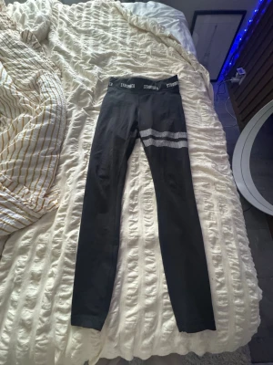 Svarta leggings från Stronger - Snygga svarta leggings från Stronger med vita prickiga ränder över låret och logga i midjan. Hög midja och stretchigt material som sitter skönt. Perfekta för träning eller chill. Lappen med storlek har tvättats bort så storleken syns ej men det är i S