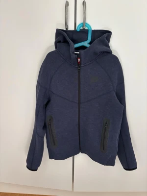Mörkblå Nike Tech hoodie - Snygg mörkblå hoodie från Nike med dragkedja framtill och två fickor med svarta dragkedjor. Tröjan har en stor huva och är tillverkad i mjukt material som passar perfekt för en avslappnad stil. Diskret Nike-logga på bröstet. Strl M junior 137-147