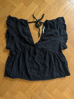 Svart croppad blus med knyt - Snygg svart croppad blus med djup v-ringning och knyt i nacken. Toppen har korta volangärmar och är tillverkad i ett lätt, strukturerat tyg som ger en avslappnad känsla. Perfekt för dig som gillar trendiga och bekväma plagg.