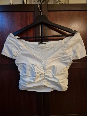 Vit croppad topp med v-ringning - Vit croppad topp med v-ringning och rynkade detaljer framtill. Toppen har korta, breda ärmar och en figurnära passform som framhäver midjan. Perfekt för en trendig och stilren outfit. Liten i storleken.