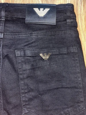 Svarta jeans från Emporio Armani - Säljer ett par svarta jeans från Emporio Armani med klassisk femficksmodell och raka ben. Jeansen har en snygg metallogo på bakfickan och patch med logga i midjan. Materialet är mjukt och stretchigt för skön passform.