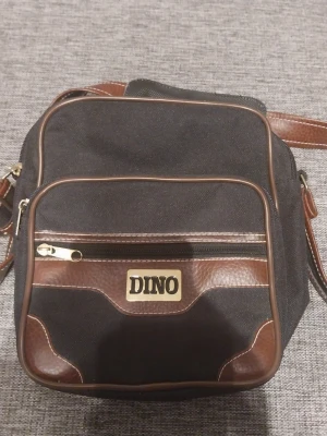 Svart axelväska med bruna detaljer DINO - Snygg svart axelväska från DINO med bruna detaljer i konstläder och guldfärgad dragkedja. Väskan har en framficka med logga, justerbar axelrem och flera fack för smart förvaring. Perfekt för dig som vill ha en stilren och praktisk väska.