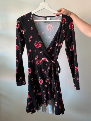 Svart blommig omlottklänning - Svart kort omlottklänning med röda och rosa blommor, långärmad och med knyt i midjan. Klänningen har v-ringad urringning och volangkant nedtill. Perfekt för vår och sommar. Använd fåtal gånger. 