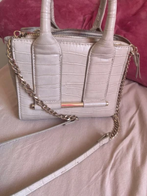 Beige croco-mönstrad handväska - Snygg beige handväska i croco-mönstrat konstläder med både handtag och avtagbar axelrem i kedja. Väskan har guldfärgade detaljer och dragkedja upptill. Perfekt storlek för det viktigaste och ger en trendig touch till din outfit.