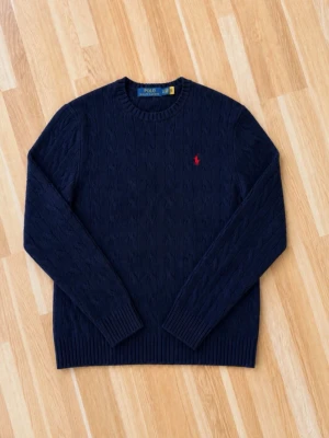 Mörkblå kabelstickad tröja Polo Ralph Lauren - Mörkblå kabelstickad tröja från Polo Ralph Lauren med rund halsringning och röd broderad logga på bröstet. Tröjan har långa ärmar och ribbade muddar vid ärmslut och nederkant. Perfekt för lager på lager och klassisk stil.
