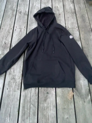 Svart hoodie från Moncler - Säljer en svart hoodie från Moncler med klassisk huva och dragsnören. Hoodien har en stor magficka framtill och Monclers ikoniska logotyp på vänster ärm. Perfekt för dig som gillar stilrena och exklusiva plagg.
