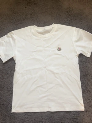 Vit t-shirt från Moncler - Säljer en stilren vit t-shirt från Moncler med klassisk rund hals och korta ärmar. På bröstet sitter Monclers ikoniska logotyp i broderad patch. Perfekt basplagg för dig som gillar exklusiva märken och enkel design.