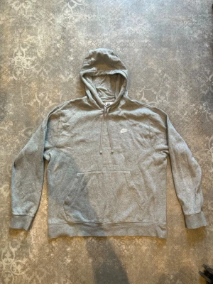 Grå hoodie från Nike med huva - Säljer en klassisk grå hoodie från Nike med broderad logga på bröstet. Hoodien har huva med dragsko och en stor magficka framtill. Tillverkad i mjukt sweatshirtmaterial och har ribbade muddar vid ärmar och nederkant.