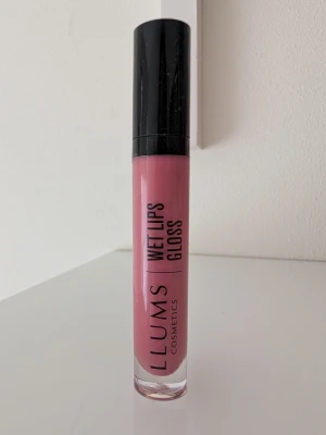 LLUMS Wet Lips Gloss 13 Happy - LLUMS Wet Lips Gloss i nyansen 13 Happy är ett läppglans med en mjuk rosa ton. Kommer i en smidig tub med svart lock och transparent kropp som visar färgen. Ger en glansig finish och är enkel att applicera med applikator. Perfekt för dig som vill ha glansiga läppar. Använd två gånger. 