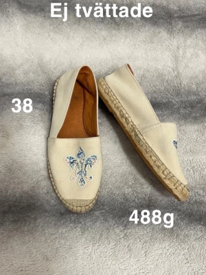 Beige loafers med broderad lilja - Säljer ett par beige loafers i tyg med broderad lilja i blått och rosa framtill. Skorna har flätad sula i jute och klassisk slip-on design. Perfekta för dig som gillar en avslappnad men ändå unik stil.🌸 Fint använt skick. Ej tvättade. Storlek 38