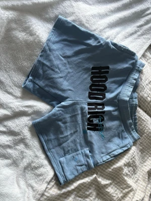 Blå HOODRICH shorts med tryck - Snygga ljusblå shorts från HOODRICH med stort svart tryck på sidan och en liten ficka framtill. Sköna att bära med elastisk midja och avslappnad .