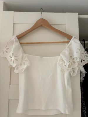 Vit topp med broderade volangärmar - Supersöt vit topp med breda, broderade volangärmar i blommigt mönster. Toppen är axelbandslös och har en lätt ribbad struktur i tyget. Perfekt för sommarens alla tillfällen.