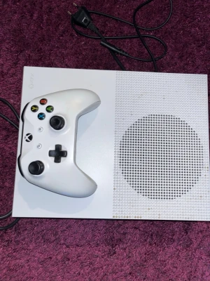 Xbox One S vit spelkonsol med handkontroll - Säljer en vit Xbox One S spelkonsol med tillhörande vit trådlös handkontroll. Konsolen har en stilren, rektangulär design med ventilerad ovansida och medföljande strömkabel. Perfekt för gaming och streaming. HDMI Kabel och Strömkabel kommer med 