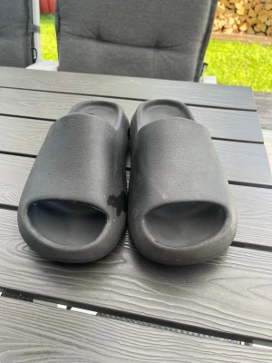 Svarta tofflor i skum från Adidas - Säljer ett par svarta tofflor från Adidas med öppen tå och chunky design. Tillverkade i mjukt och lätt skummaterial för maximal komfort. Perfekta att glida i hemma eller ute på sommaren. Adidas-logga syns på innersulan. (Storlek: 42/43)