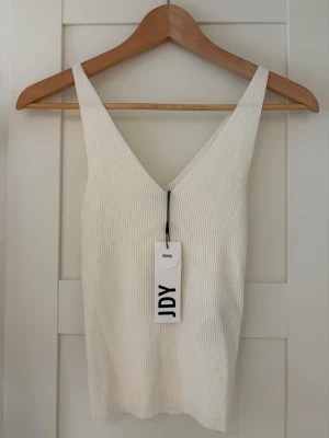 Vit ribbad linnetopp från JDY - Snygg vit ribbad linnetopp från JDY med v-ringning både fram och bak. Toppen är ärmlös och har en smickrande slim passform, perfekt till sommaren. Enkel att matcha med både jeans och kjol.