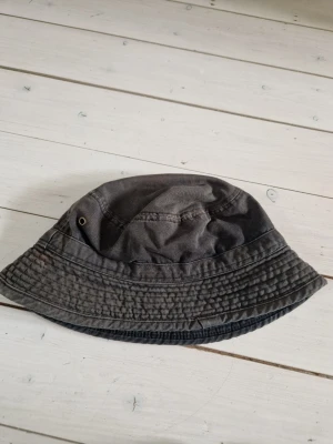 Mörkgrå bucket hat i bomull - En stilren mörkgrå bucket hat i mjuk bomull med klassisk form och sydda detaljer runt brättet. Hatten har metallöljetter på sidorna för ventilation och passar perfekt för en avslappnad streetwear-look.
