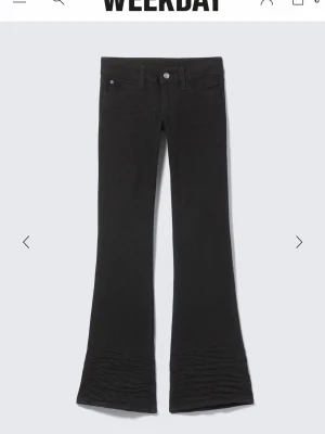 Svarta bootcut jeans från Weekday - Säljer ett par svarta bootcut jeans från Weekday med klassisk femficksmodell och utsvängda ben. Jeansen har en knapp och dragkedja framtill samt en lila patch bak på linningen. Perfekta för dig som gillar en retroinspirerad look.