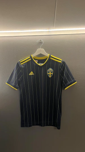 Svensk fotbollströja Adidas blå/gul - Mörkblå fotbollströja från Adidas med tunna vita ränder, gula detaljer på axlar och ärmslut samt Sveriges landslagsemblem på bröstet. Tillverkad i lätt och ventilerande material, perfekt för träning eller match.