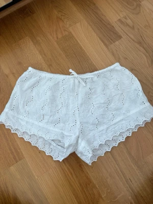 Vita broderade shorts med spetskant - Säljer ett par vita shorts med broderade detaljer och vågig spetskant. Shortsen har snörning i midjan och är tillverkade i ett lätt bomullstyg, perfekta för varma dagar. Storlek 146/152