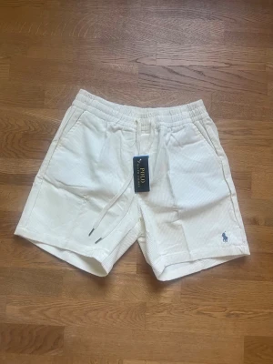 Vita manchester shorts från Polo Ralph Lauren - Säljer ett par vita manchester shorts från Polo Ralph Lauren med elastisk midja och snörning. Klassisk passform med fickor på sidorna och en liten broderad logga i blått framtill. Perfekta för en avslappnad och stilren look.