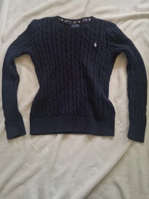 Mörkblå kabelstickad tröja Polo Ralph Lauren - Kabelstickad mörkblå tröja från Polo Ralph Lauren med rund halsringning och lång ärm. Tröjan har den klassiska broderade loggan på bröstet och ribbstickade muddar vid ärmslut och nederkant. Perfekt för en stilren och tidlös look 💕 storlek XL i barnstorlek så motsvarande xs skulle jag tro! Be om bilder på!