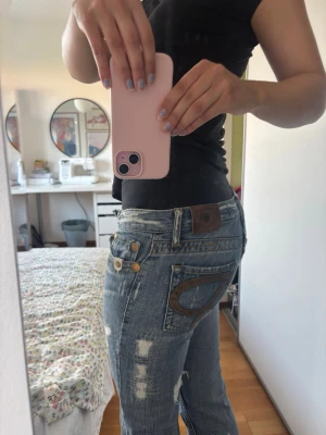 Blå slitna jeans med detaljer - Säljer ett par blå jeans med slitningar och unika detaljer på bakfickan. Jeansen har raka ben som är utsvängda där nere, låg midja och dekorativa nitar vid fickorna. Materialet är klassisk denim i bomull och de har en avslappnad passform.