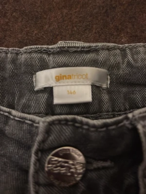 Grå jeans från Gina Tricot med detaljer - Snygga grå jeans från Gina Tricot i storlek 146. Jeansen har coola fickdetaljer med flätad söm och knappstängning. Perfekta för dig som vill ha en unik look med extra detaljer. Klassisk femficksmodell och mjukt denimtyg. Den har en knapp som åkt av på ena fickan