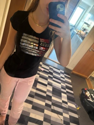 Svart GUESS t-shirt med paljetter - Snygg svart t-shirt från GUESS med korta ärmar och glittriga paljetter i silver, rött och svart på bröstet. T-shirten har en v-ringad hals och en normal passform. Perfekt för dig som vill sticka ut lite extra.