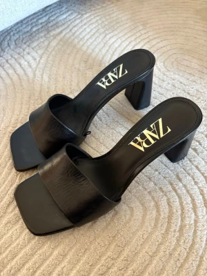 Svarta klackpumps från Zara - Stilrena svarta pumps från Zara med bred rem över foten och fyrkantig tå. Skorna har en tjockare klack och är tillverkade i ett glansigt material som liknar läder. Använda enstaka gånger och är i fint skick. 