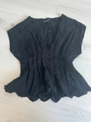 Svart broderad blus från Gina Tricot - Snygg svart blus från Gina Tricot med broderade detaljer och v-ringning. Blusen har korta ärmar och elastisk midja som ger en fin siluett. Materialet är lätt och har hålbroderier längs kanterna för en romantisk touch. Superfin på!