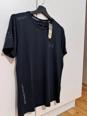 Svart Under Armour t-shirt - Snygg svart t-shirt från Under Armour med diskret logga på bröstet och tryck på ärmen samt sidan. Tillverkad i ett lätt och mjukt syntetmaterial som andas, perfekt för träning. Klassisk rund hals och korta ärmar för en sportig look.