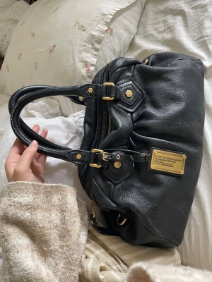 Svart handväska med gulddetaljer - Trendig berömd Marc Jacobs väska med båda axelband och används främst som handväska, lyxig och så bra utrymme, får plats med mycket mer än vad man tror. Den är praktisk och passar både till skolan och till andra möten älskar!!🌸🌸 Lite effekter men inget man tänker på.