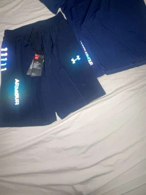 Mörkblå träningsshorts med tryck och tröja - Säljer ett par mörkblå träningsshorts med resårmidja och snörning. Shortsen har färgglada tryckdetaljer och logga på benet. Perfekta för träning eller sportiga dagar. Materialet känns lätt och andas bra. Det helt ny storlek mellan S och M båda passar