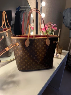 Monogram väska - Säljer en handväska i monogrammönster med röd insida och  med guldiga detaljer. Väskan kommer med en matchande liten clutch. Väskan är i nyskick, kommer även i påsen den kom i!💛