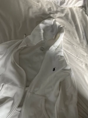 Vit hoodie från Polo Ralph Lauren - Säljer en vit hoodie från Polo Ralph Lauren med dragkedja framtill och klassisk logga broderad på bröstet. Tröjan har huva med dragsko och två fickor framtill. Perfekt för en clean och stilren look.