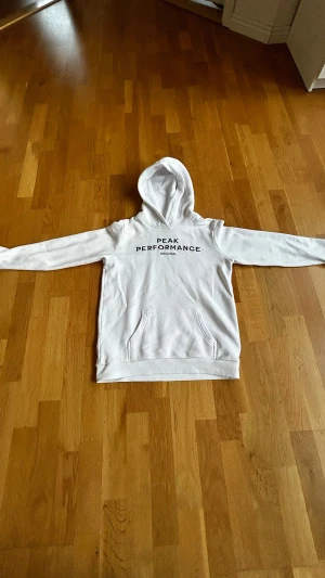 Vit Peak Performance hoodie - Snygg vit hoodie från Peak Performance med svart text på bröstet. Klassisk modell med huva och stor magficka framtill. Tillverkad i mjukt material och har ribbade muddar vid ärmslut och nederkant. Perfekt för en avslappnad och sportig stil. Hör av er vid minsta fundering 😁