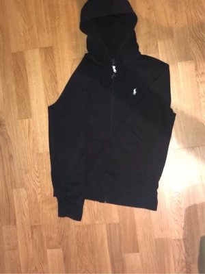 Svart hoodie med dragkedja - Svart hoodie med dragkedja och huva. Klassisk design med fickor framtill och justerbara snören i huvan. Liten vit logga broderad på bröstet. Perfekt för en avslappnad och stilren look.